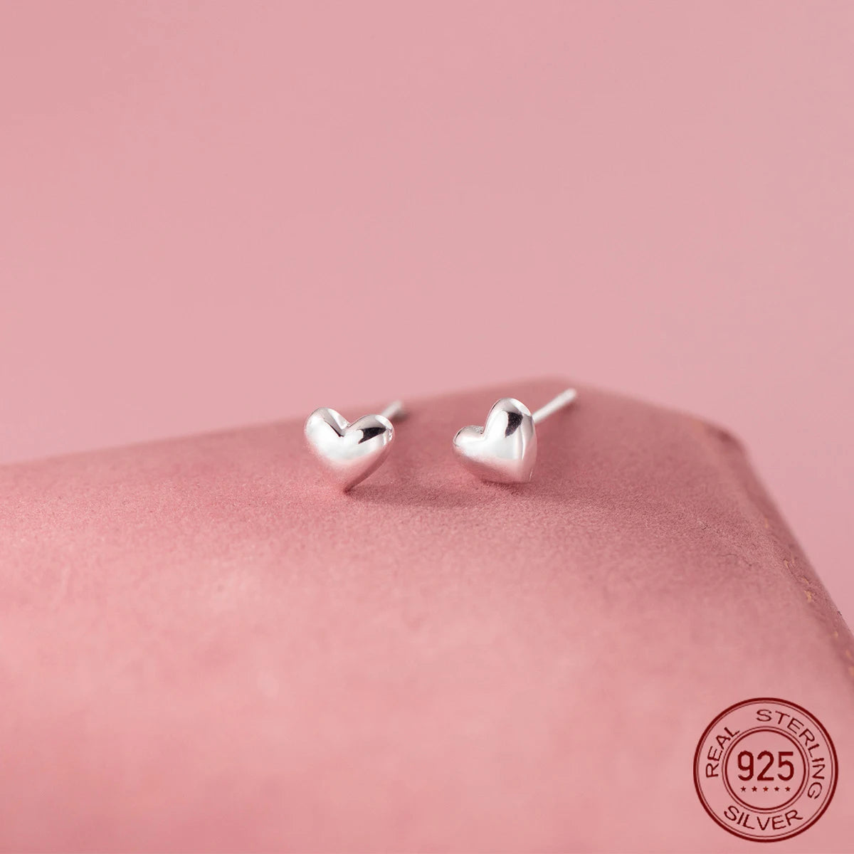 Kleine Herz-Ohrstecker aus poliertem 925 Sterling Silber mit leicht gewölbter 3D-Optik auf rosafarbenem Untergrund.
