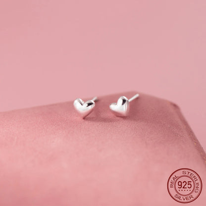 Kleine Herz-Ohrstecker aus poliertem 925 Sterling Silber mit leicht gewölbter 3D-Optik auf rosafarbenem Untergrund.