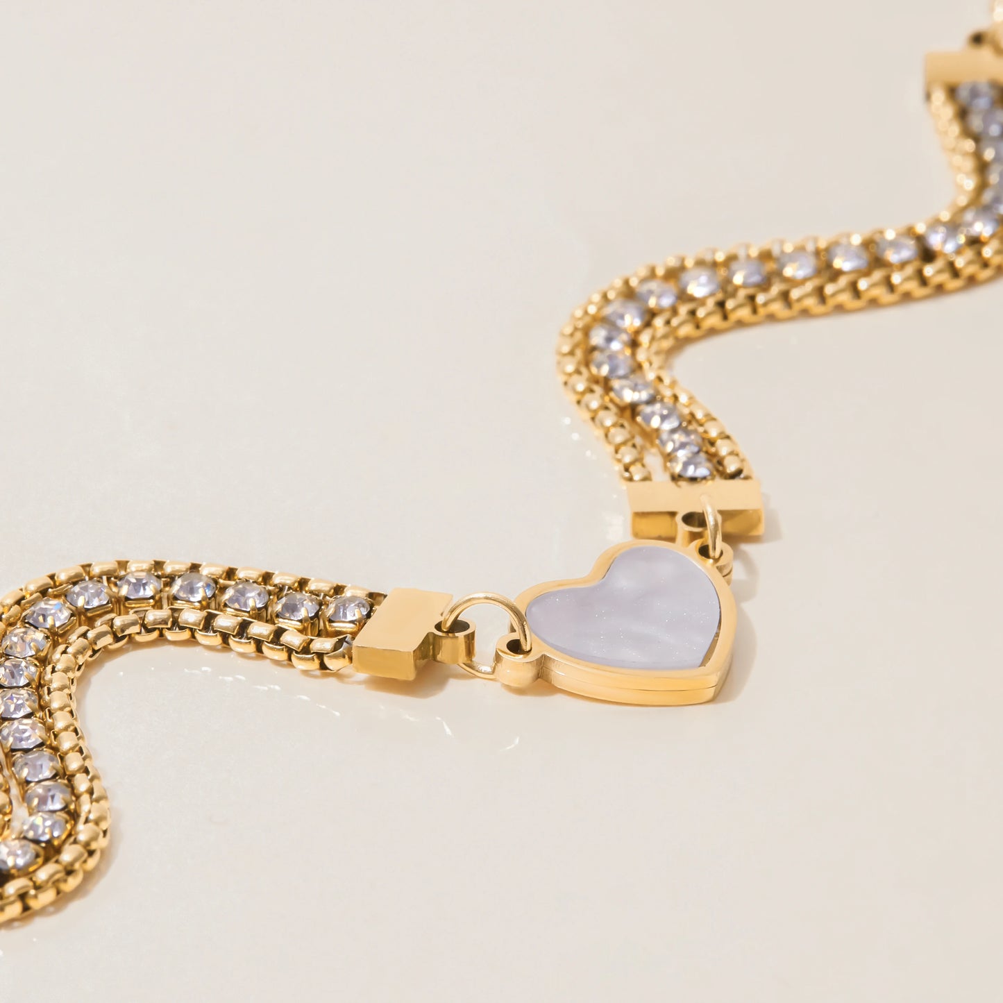 Ein dreireihiges, goldfarbenes Gliederarmband mit funkelnden Zirkoniasteinen und einem zentralen Herz, wellenförmig auf hellem Untergrund präsentiert.