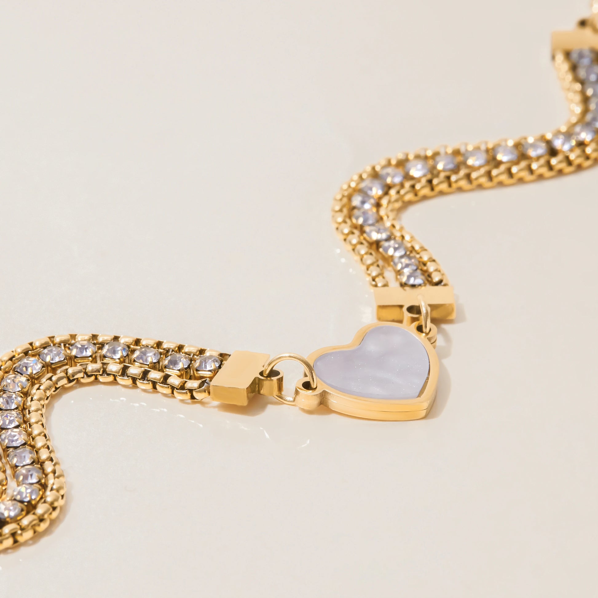 Ein dreireihiges, goldfarbenes Gliederarmband mit funkelnden Zirkoniasteinen und einem zentralen Herz, wellenförmig auf hellem Untergrund präsentiert.