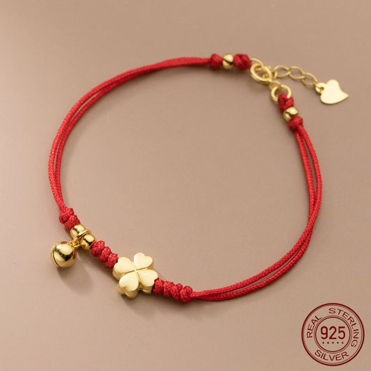Rotes Textilarmband aus 925er Sterling Silber mit 18K Vergoldung, verziert mit einem vierblättrigen Kleeblatt und einem kleinen Glöckchen auf beigem Untergrund.