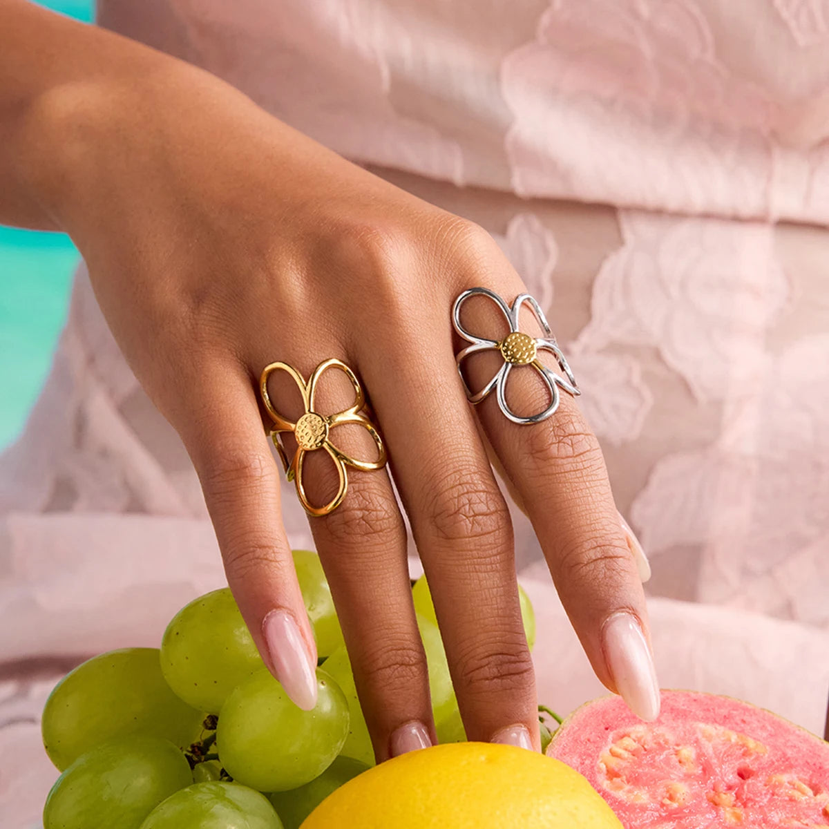 Nahaufnahme einer Hand, die einen großen, goldenen Ring mit einem Blumen-Design trägt. Die Blätter sind aus Metallschleifen geformt. Der Ring ist auf einem hellen Hintergrund mit Früchten wie Weintrauben und einer halbierten Guave abgebildet.