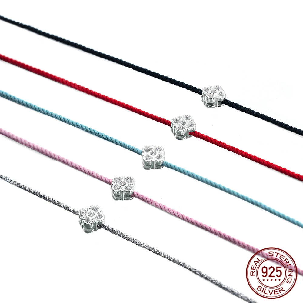 Fünf filigrane Armbänder mit farbigen Textilbändern (Schwarz, Rot, Hellblau, Rosa und Silberfarben), die nebeneinander auf weißem Hintergrund liegen. Jedes Armband hat einen kleinen, zentralen Anhänger in Blüten- oder Kleeblattform aus 925er Sterling Silber, der mit Zirkoniasteinen besetzt ist. Das 925er Sterling Silver Echtheitssiegel ist in der unteren rechten Ecke sichtbar.