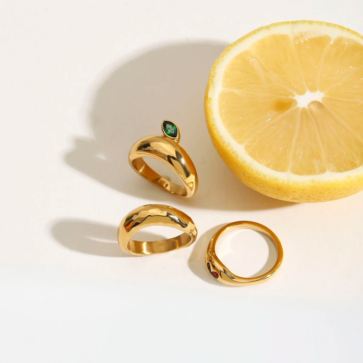 Drei Ringe aus 18K vergoldetem Edelstahl, ein Schlangenring, ein Ring mit grünem Stein und ein Herz-Ring, neben einer Zitrone.