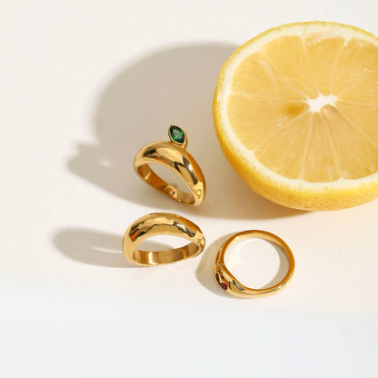 Drei Ringe aus 18K vergoldetem Edelstahl, ein Schlangenring, ein Ring mit grünem Stein und ein Herz-Ring, neben einer Zitrone.