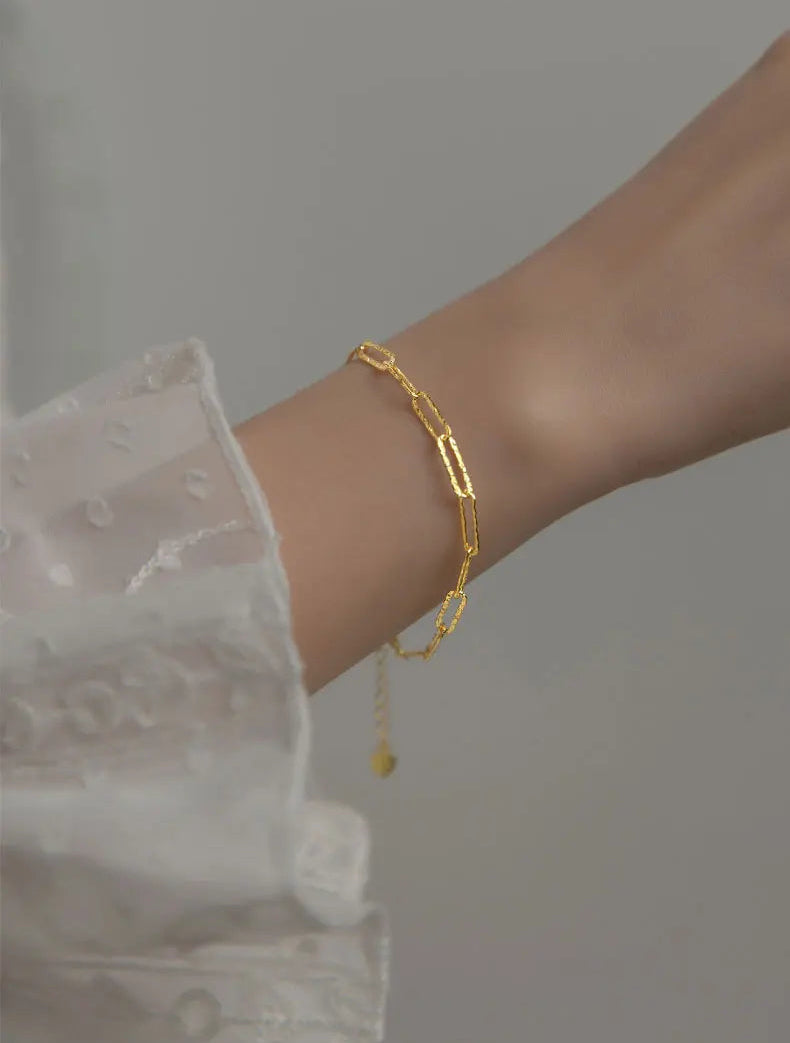 Handgelenk, das ein zartes, goldfarbenes Armband trägt (alternatives Modell oder Farbvariante), kombiniert mit einem langärmeligen, weißen Oberteil mit Rüschen.