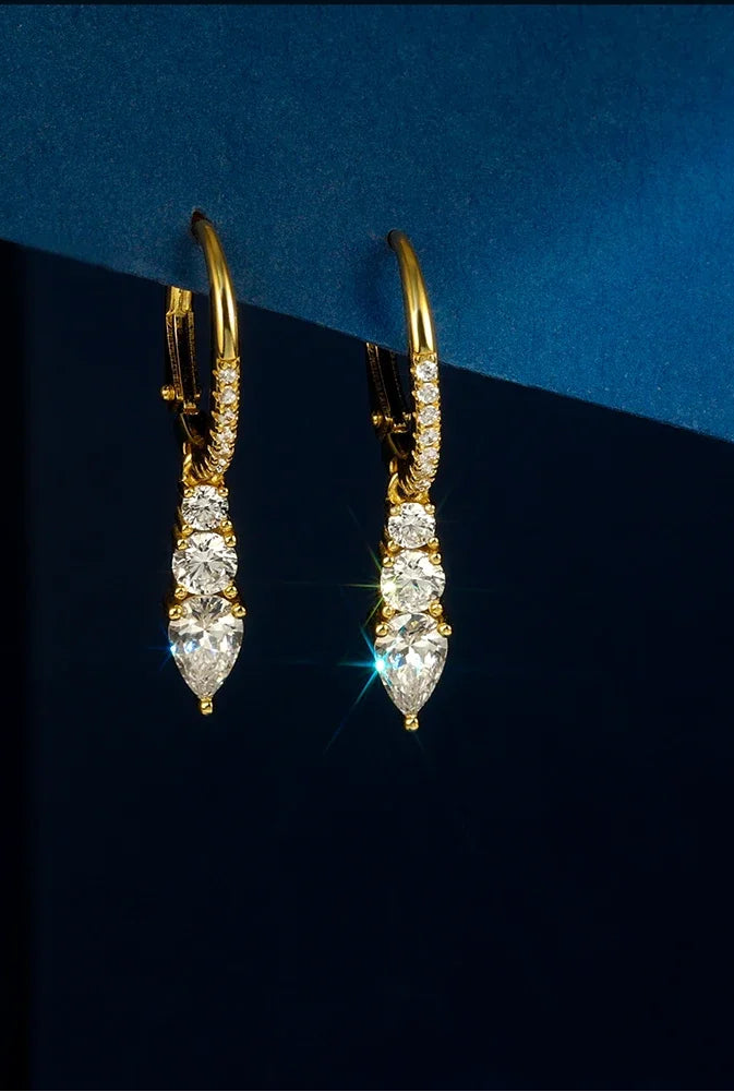 Ein Paar goldener, hängender Ohrringe aus vergoldetem 925er Silber, jeweils besetzt mit drei länglichen Zirkonia-Steinen, präsentiert auf dunklem Hintergrund.
