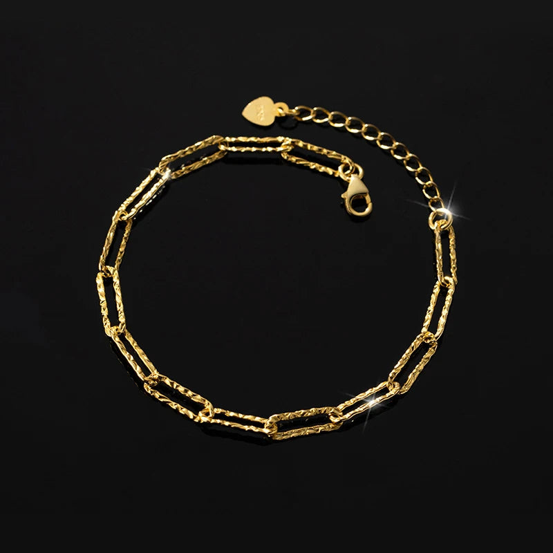 Goldfarbenes Kettenarmband im "Paperclip"-Stil mit länglichen, unregelmäßig gehämmerten Gliedern. Es hat einen Karabinerverschluss und eine Verlängerungskette, die mit einem kleinen Herz-Anhänger endet.