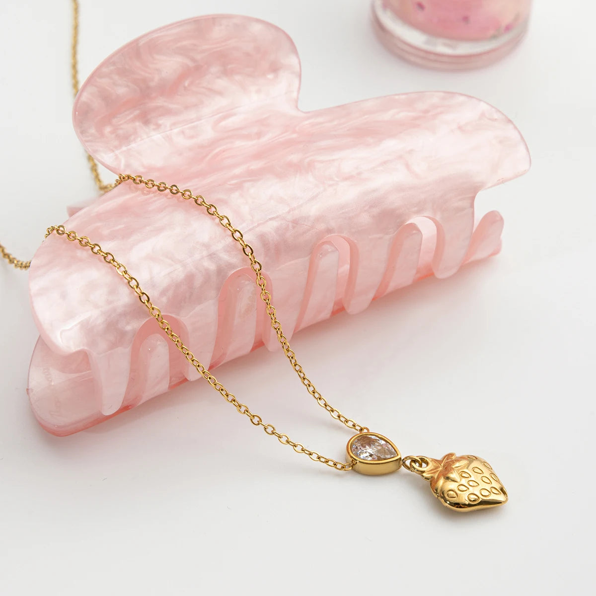 Goldene Halskette aus Edelstahl mit 18K Vergoldung, funkelndem Zirkonia-Stein und strukturiertem Herz, dekorativ arrangiert neben einer rosa Haarklammer aus Acryl.