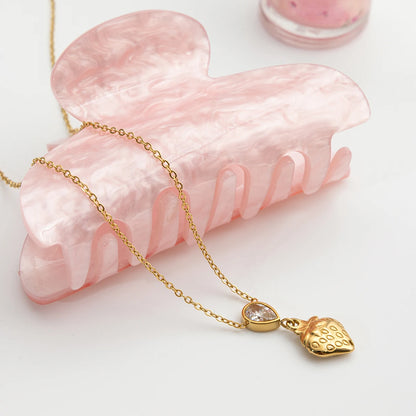 Goldene Halskette aus Edelstahl mit 18K Vergoldung, funkelndem Zirkonia-Stein und strukturiertem Herz, dekorativ arrangiert neben einer rosa Haarklammer aus Acryl.
