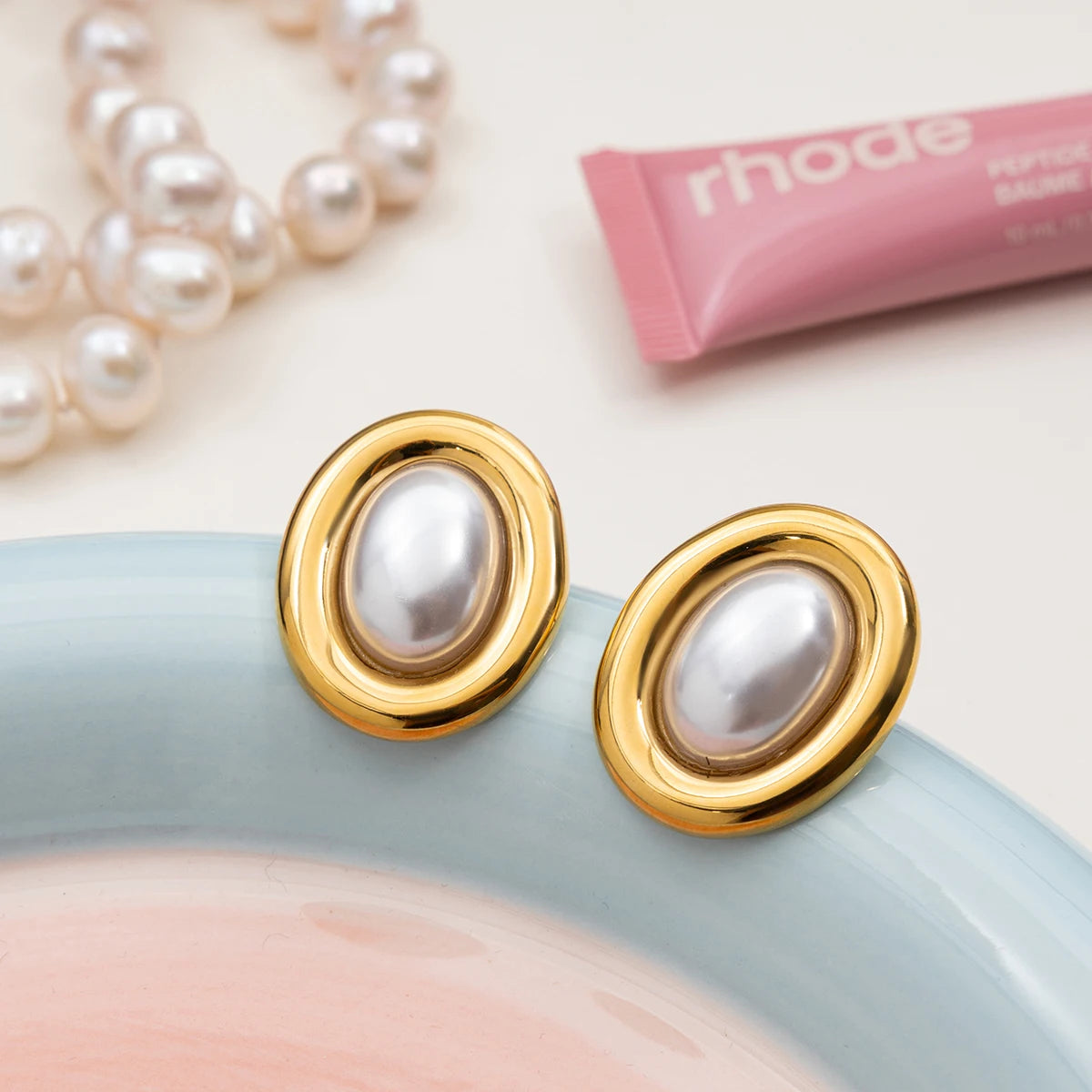 Zwei goldene Ohrstecker mit weißen Harzperlen liegen auf einem pastellfarbenen Teller, daneben ein rosafarbener Lippenstift. Eine schicke Schmuckszene für einen Beauty-orientierten Kontext.