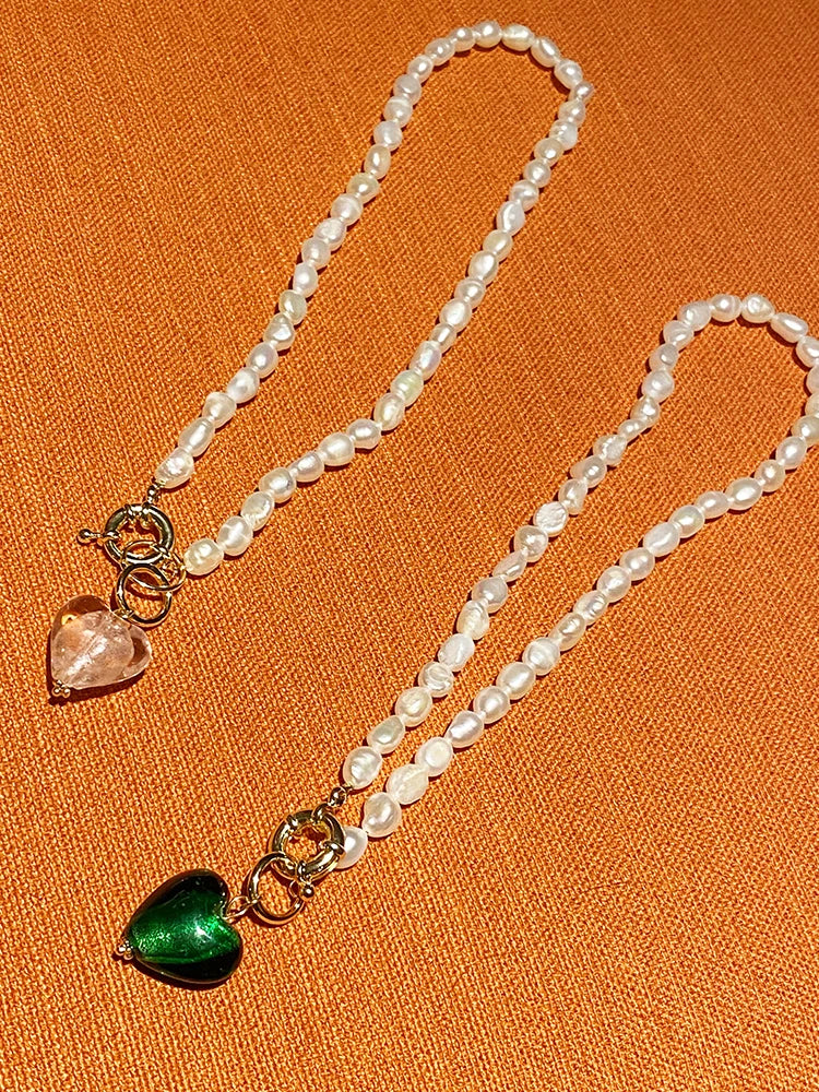 Zwei Naturperlenketten mit vergoldeten Titan-Verschlüssen und Herz-Anhängern in Rosa und Grün auf orangefarbenem Grund.
