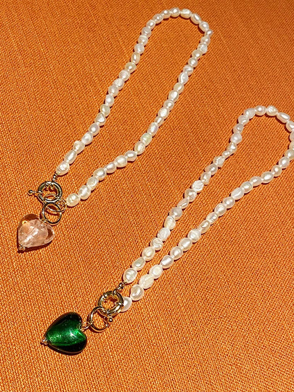 Zwei Naturperlenketten mit vergoldeten Titan-Verschlüssen und Herz-Anhängern in Rosa und Grün auf orangefarbenem Grund.