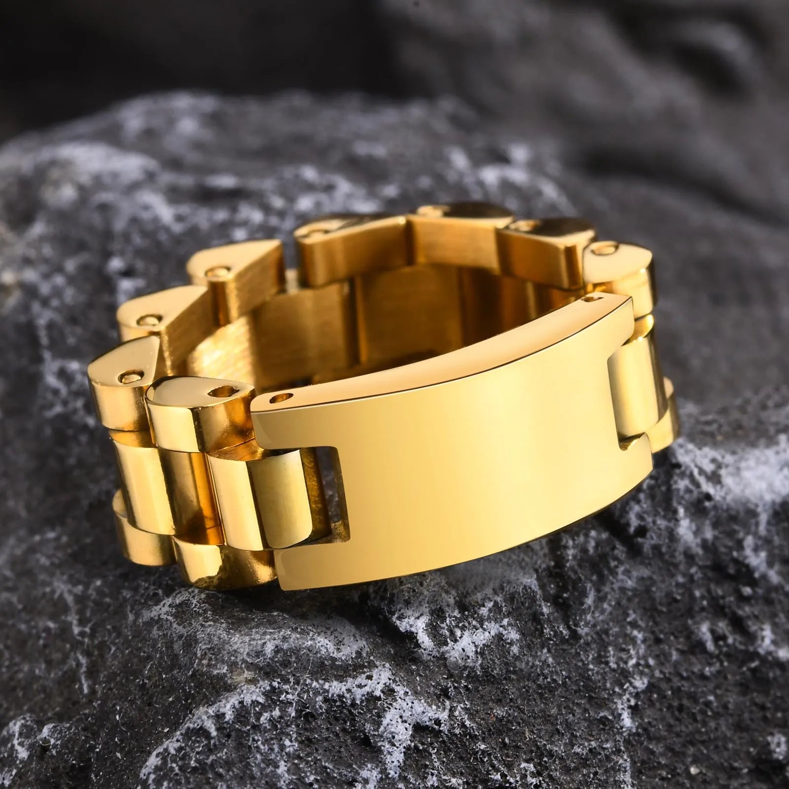 Freigestellte Detailansicht eines breiten, goldfarbenen Edelstahl-Rings mit Platten- und Glieder-Muster, liegend auf einem dunklen, steinernen Untergrund.