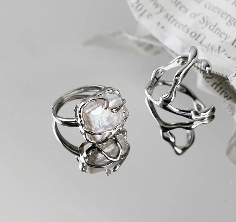 Zwei abstrakte, offene Ringe aus 925er Silber auf einer spiegelnden Oberfläche, wobei einer einen unregelmäßigen weißen Stein trägt und der andere ein skulpturales, unebenes Design aufweist.

