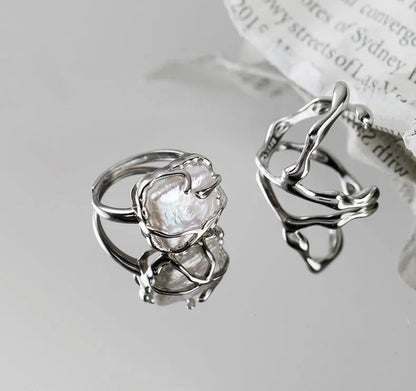 Zwei abstrakte, offene Ringe aus 925er Silber auf einer spiegelnden Oberfläche, wobei einer einen unregelmäßigen weißen Stein trägt und der andere ein skulpturales, unebenes Design aufweist.

