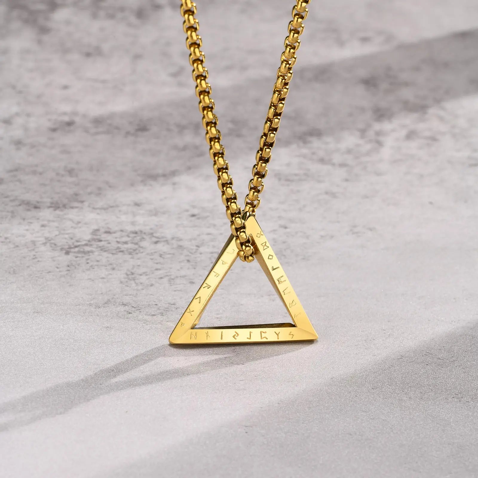 Nahaufnahme der goldfarbenen Edelstahlkette mit dreieckigem Anhänger, wobei die Gravur/Prägung auf dem unteren Rand des Dreiecks sichtbar ist, auf hellem Hintergrund.
