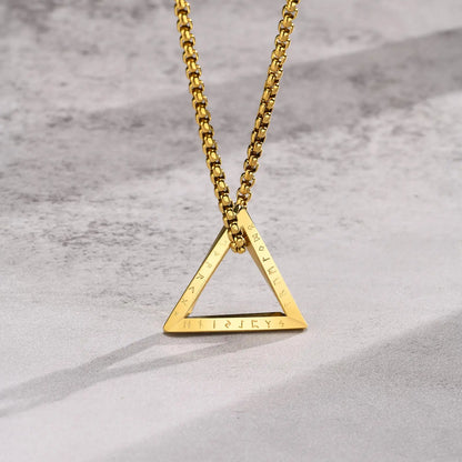 Nahaufnahme der goldfarbenen Edelstahlkette mit dreieckigem Anhänger, wobei die Gravur/Prägung auf dem unteren Rand des Dreiecks sichtbar ist, auf hellem Hintergrund.