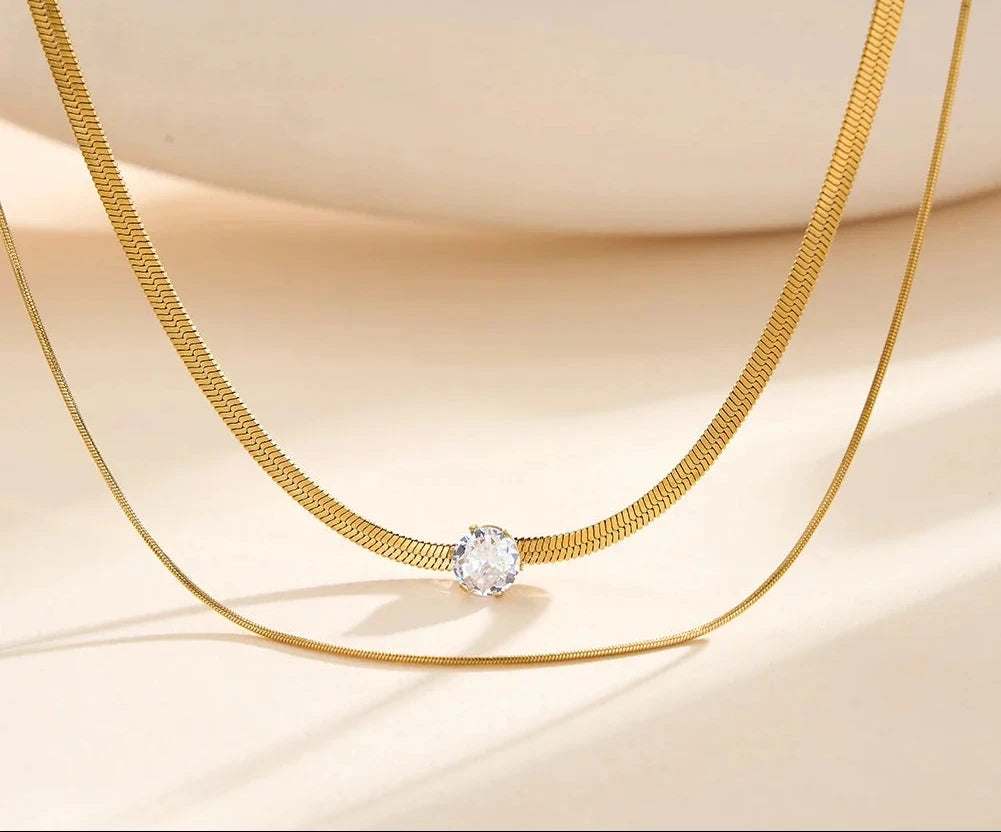Nahaufnahme der 18K vergoldeten, zweireihigen Edelstahl-Kette mit funkelndem Zirkonia-Anhänger vor hellem Hintergrund.