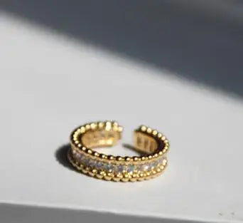 Nahaufnahme eines vergoldeten Rings auf weißem Grund, die das offene Design und die dreireihige Optik zeigt: zwei äußere Reihen aus Goldperlen-Struktur und eine mittlere Reihe aus glitzernden Zirkonia.