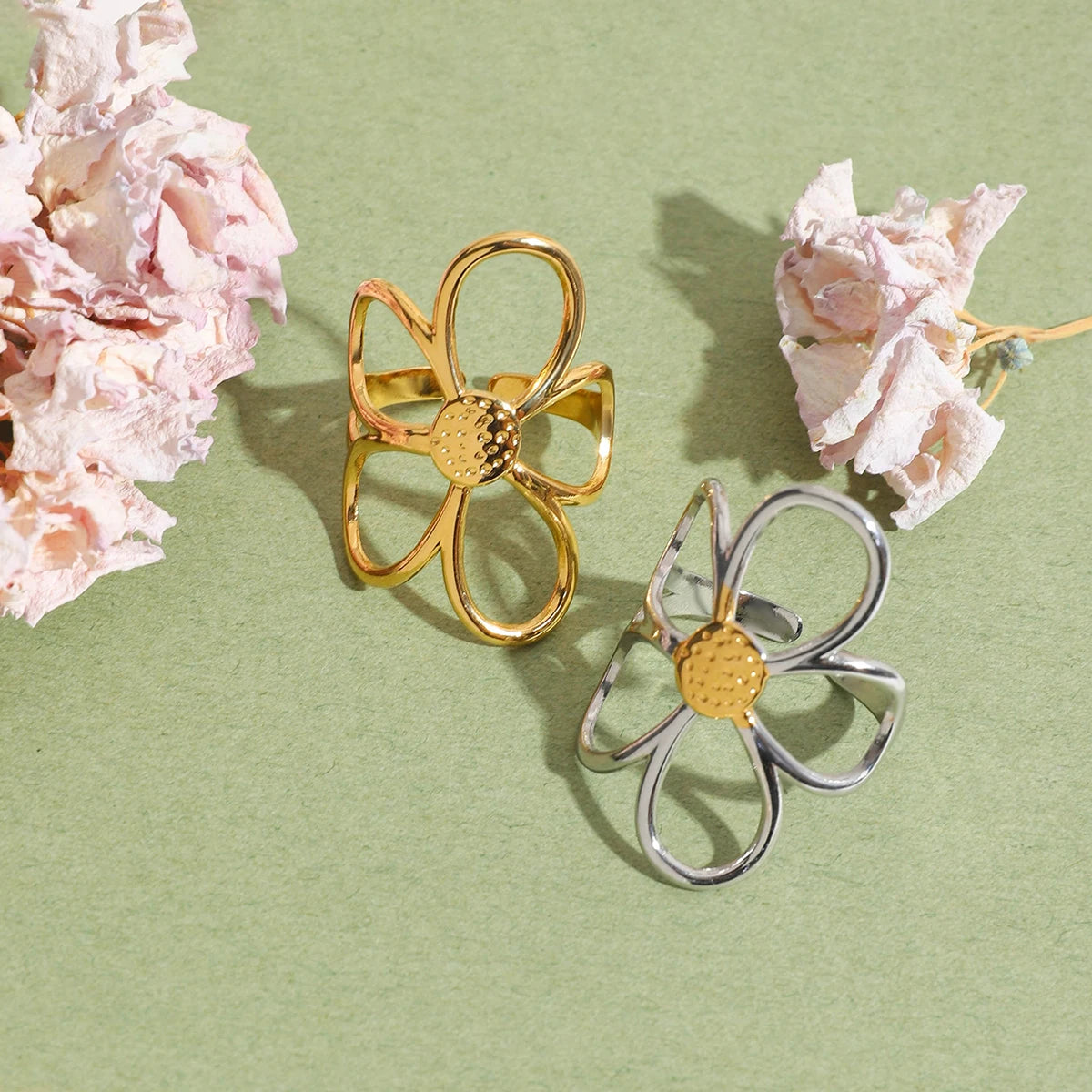 Die goldenen und silbernen Versionen des Rings mit Blumen-Design liegen nebeneinander. Die goldene Variante hat einen metallenen Schleifenlook, die silberne eine glatte Oberfläche. Im Hintergrund sind Blumen und Blätter zu sehen.