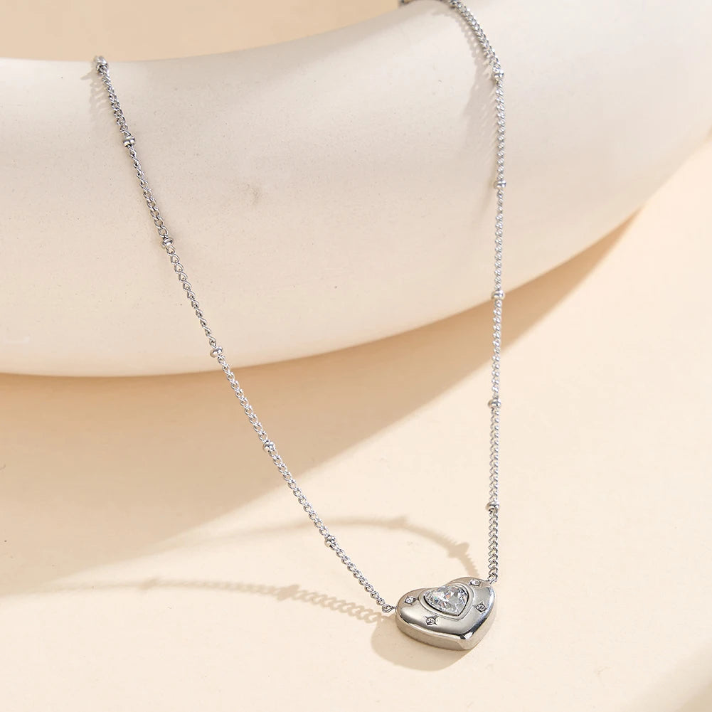 Silberne Edelstahl-Halskette mit Zirkonia-Herzanhänger elegant auf hellem Dekorationsstein präsentiert.