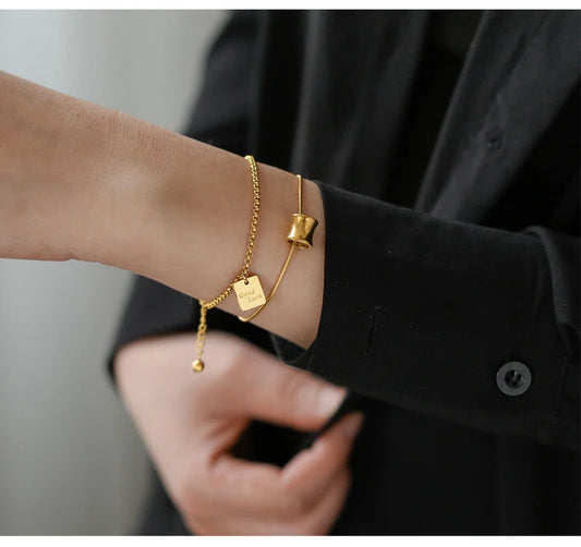 Tragestyling: Zwei 18K vergoldete Titan-Armbänder im Layering-Look, eines mit Zylinder-Anhänger und eines mit Plättchen.