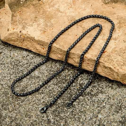 Lange, schwarze Edelstahl-Kette im Vintage-Stil mit einem dicken Zopfmuster (Weizenmuster), ausgebreitet auf einem rauen, steinernen Hintergrund. Der Karabinerverschluss ist sichtbar.