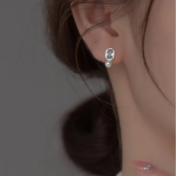 Seitenprofil einer Frau, die einen filigranen Ohrstecker aus Silber mit blauem Zirkonia-Edelstein und hängender Perle am Ohrläppchen trägt.