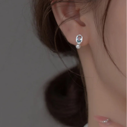 Seitenprofil einer Frau, die einen filigranen Ohrstecker aus Silber mit blauem Zirkonia-Edelstein und hängender Perle am Ohrläppchen trägt.