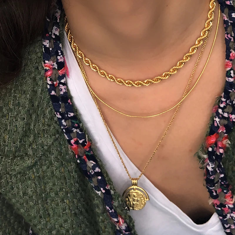 Detaillierte Nahaufnahme einer Frau, die mehrere goldene Halsketten trägt, darunter eine dicke Kordelkette und ein Medaillon-Anhänger.