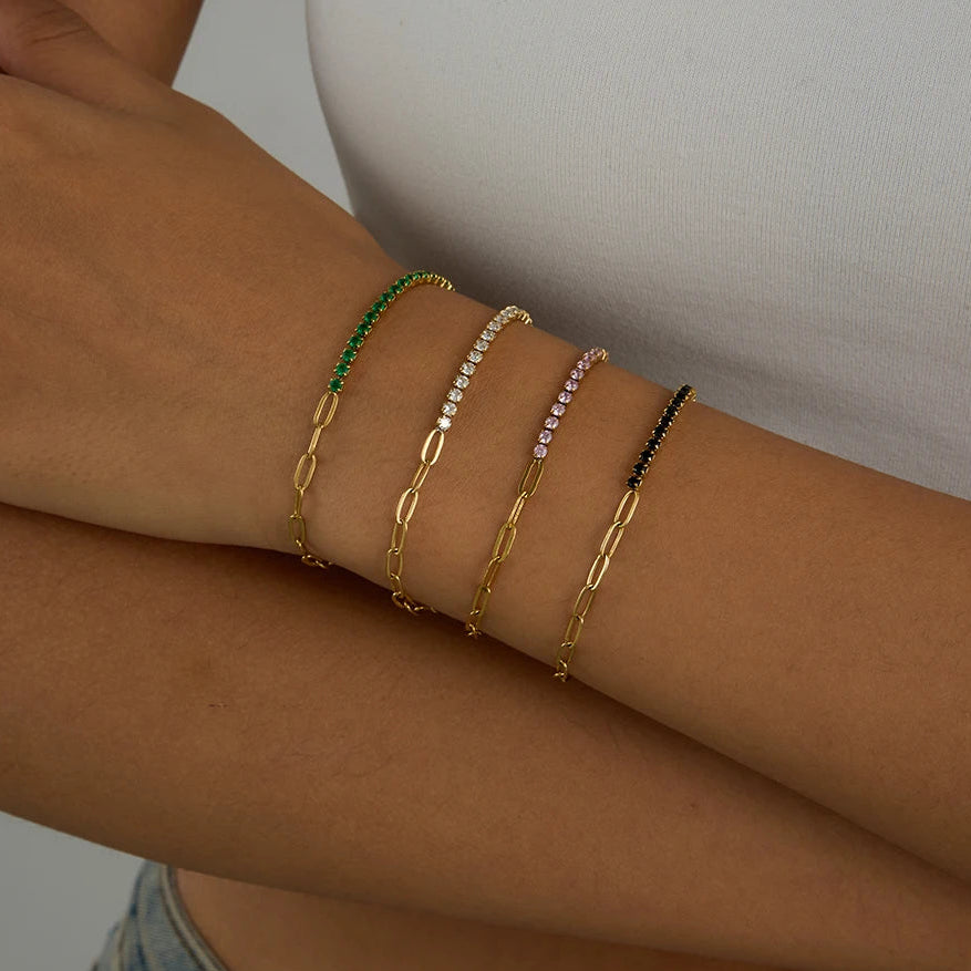 „Vier 18K vergoldete Edelstahl-Armbänder im modischen Layering-Look am Handgelenk, besetzt mit funkelnden Zirkonia-Steinen in Grün, Weiß, Rosa und Schwarz.“
