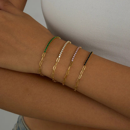 „Vier 18K vergoldete Edelstahl-Armbänder im modischen Layering-Look am Handgelenk, besetzt mit funkelnden Zirkonia-Steinen in Grün, Weiß, Rosa und Schwarz.“