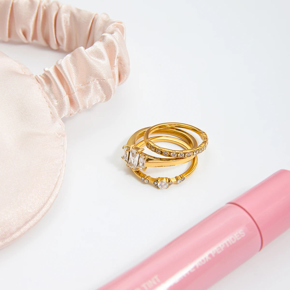 Zwei goldene Ringe mit Zirkoniasteinen liegen neben einer pinken Schlafmaske und einem Wimperntuschef-Produkt, was ein Beauty- und Wellness-Thema aufgreift.