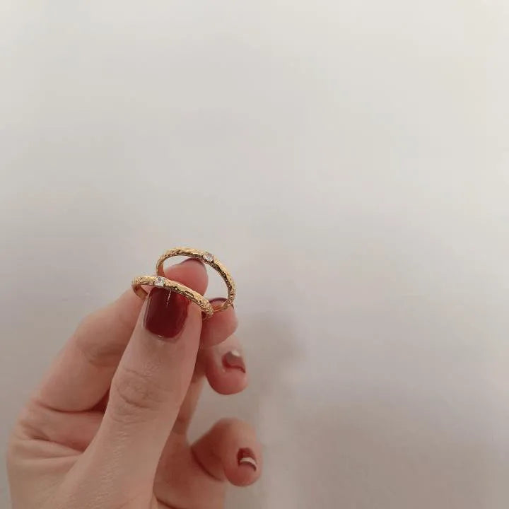 Zwei filigrane, vergoldete Ringe mit gehämmerter Oberfläche und Zirkonia, gehalten zwischen zwei Fingern.