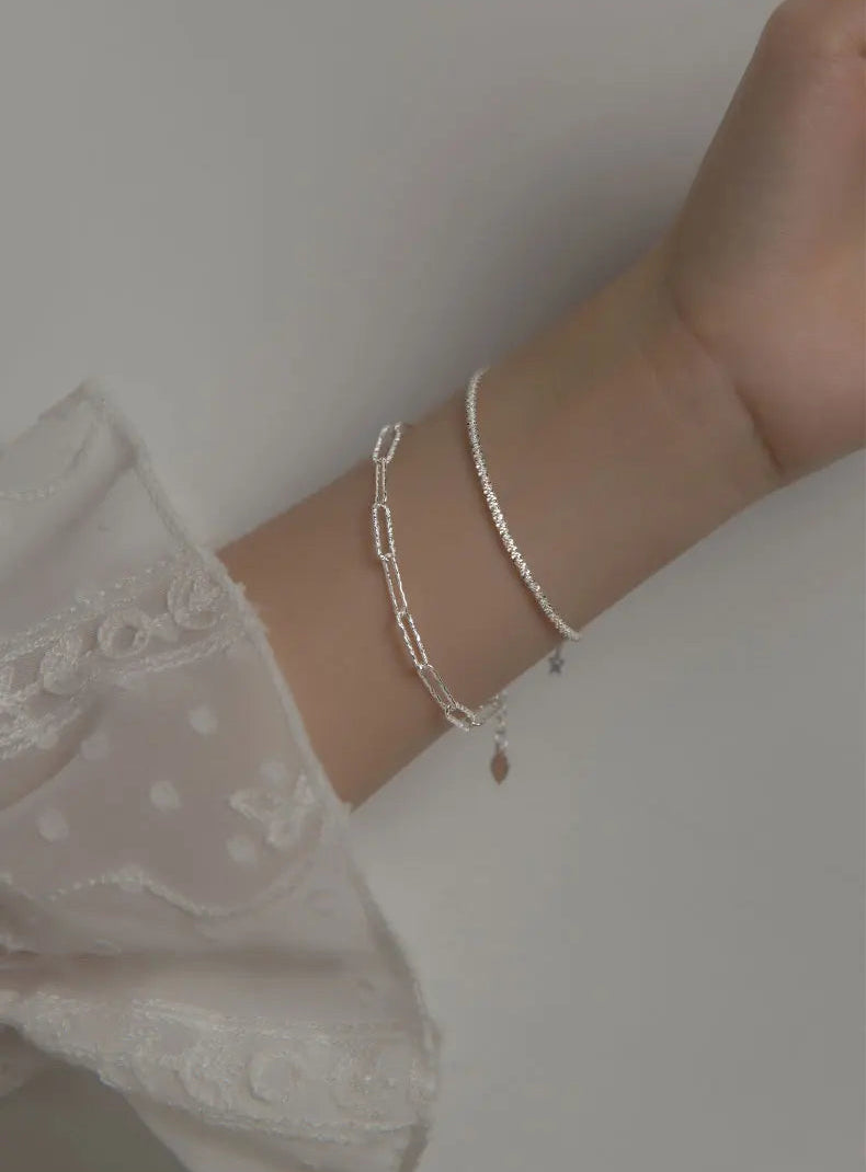 Handgelenk, das zwei zarte, silberfarbene Armbänder trägt. Eines ist ein dünnes Kettchen, das andere ein längliches Gliederketten-Armband.