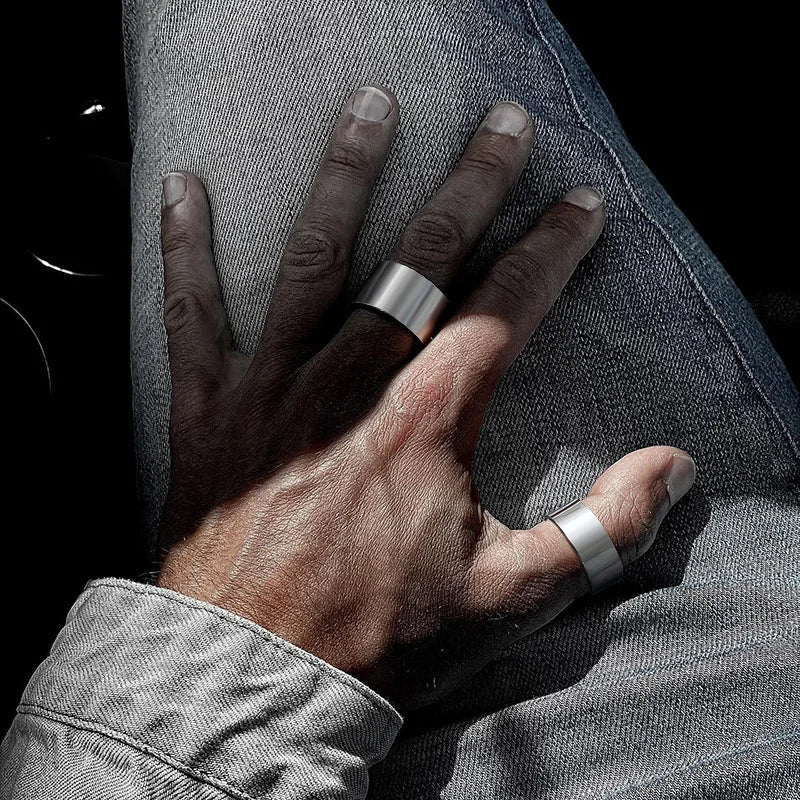 Nahaufnahme einer Männerhand in einer Jeansjacke: Ein breiter, offener Edelstahl-Bandring wird am Mittelfinger und ein weiterer am Daumen getragen.