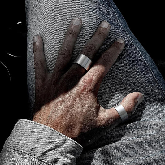 Nahaufnahme einer Männerhand in einer Jeansjacke: Ein breiter, offener Edelstahl-Bandring wird am Mittelfinger und ein weiterer am Daumen getragen.