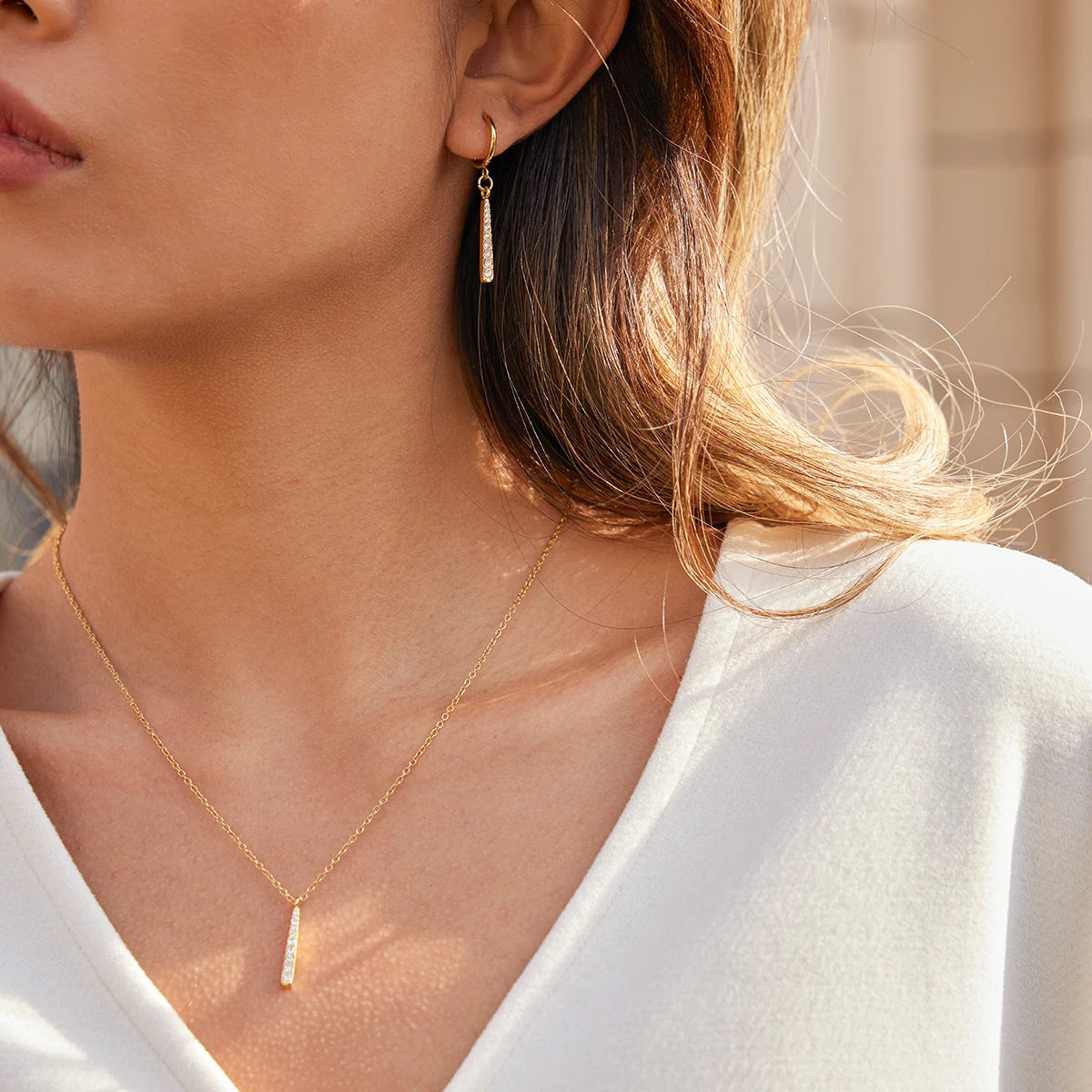 Eine Frau trägt ein elegantes Schmuckset aus goldbeschichtetem Edelstahl (PVD); die Halskette und die Ohrringe verfügen über längliche Anhänger, die mit funkelnden Strasssteinen besetzt sind.