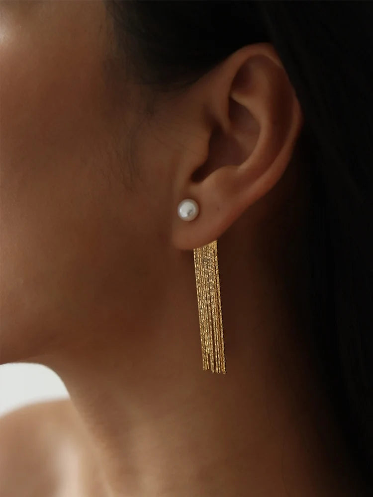Seitliche Nahansicht einer Frau, die einen eleganten Ohrring trägt; eine weiße Süßwasserperle sitzt auf dem Ohrläppchen, von der ein langer, fächerartiger Besatz aus feinen, 14k vergoldeten Messingketten herabhängt.