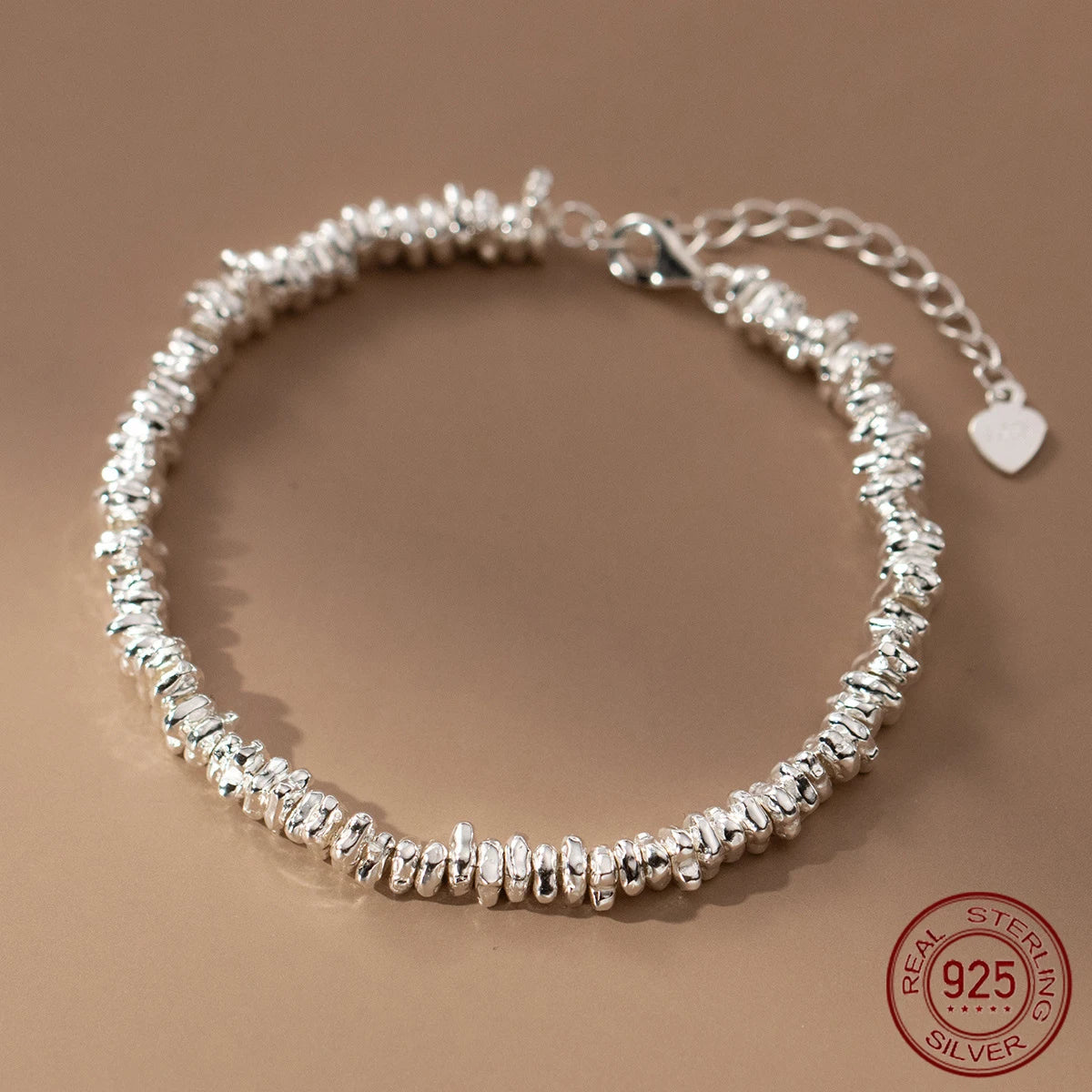  Detailaufnahme eines silberfarbenen Armbands aus 925er Sterling Silber, das aus unregelmäßig geformten, "zerknüllten" Elementen besteht und einen verstellbaren Kettenverschluss mit einem kleinen Herz-Anhänger aufweist. Das 925er Siegel ist sichtbar.