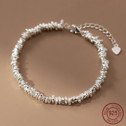  Detailaufnahme eines silberfarbenen Armbands aus 925er Sterling Silber, das aus unregelmäßig geformten, "zerknüllten" Elementen besteht und einen verstellbaren Kettenverschluss mit einem kleinen Herz-Anhänger aufweist. Das 925er Siegel ist sichtbar.