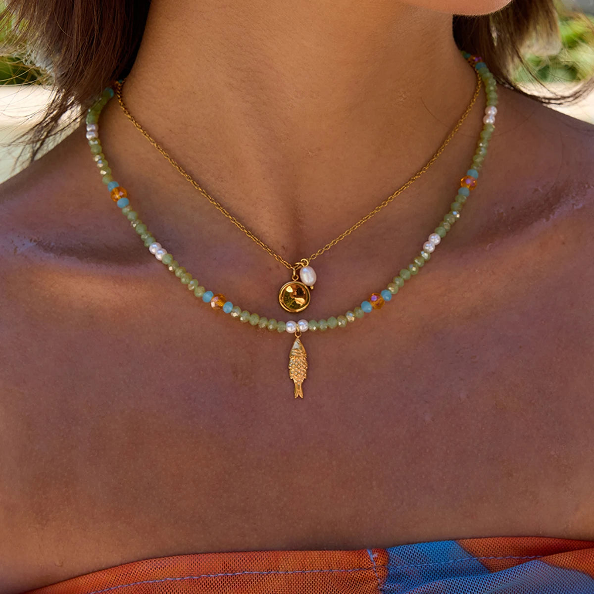 Zweireihiger Layering-Look am Dekolleté: Eine feine goldene Edelstahlkette mit Plättchen-Anhänger und Süßwasserperle neben einer bunten Perlenkette mit echten Perlen und goldenem Fisch.