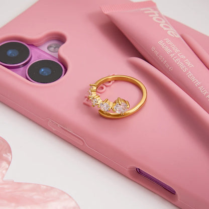 „Moderner Ring aus 18K vergoldetem Edelstahl mit Zirkonia-Steinen, platziert auf einer rosa Smartphone-Hülle neben einer Tube Lipgloss.“