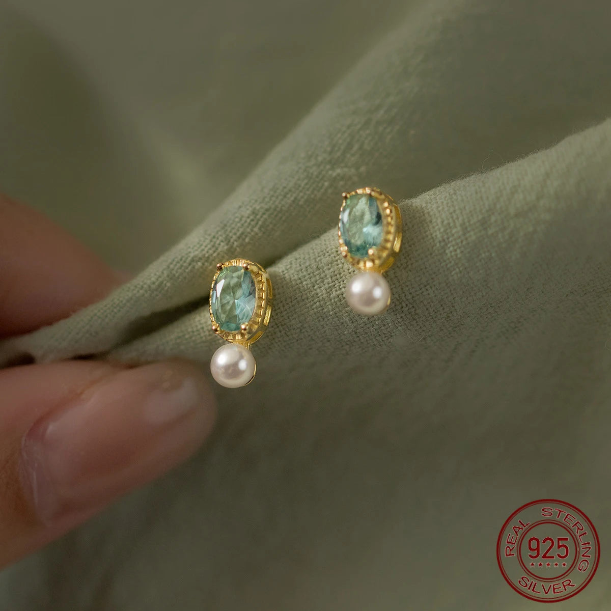 Nahaufnahme von eleganten Ohrringen aus 18K vergoldetem 925er Sterling Silber, besetzt mit einem ovalen, hellblauen Zirkonia-Stein und einer hängenden weißen Perle auf grünem Stoff.