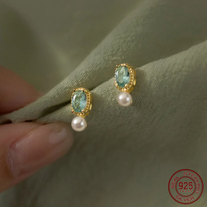 Nahaufnahme von eleganten Ohrringen aus 18K vergoldetem 925er Sterling Silber, besetzt mit einem ovalen, hellblauen Zirkonia-Stein und einer hängenden weißen Perle auf grünem Stoff.