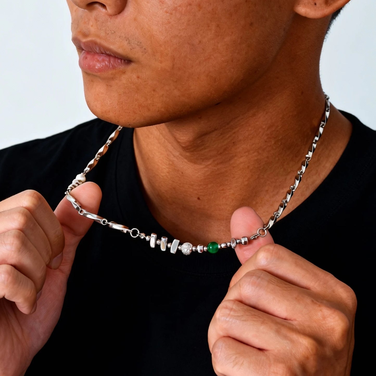 Nahaufnahme eines Mannes, der eine asymmetrische Edelstahlkette mit integrierten Metallperlen, einer weißen Süßwasserperle und einer grünen Jade-Perle trägt.
