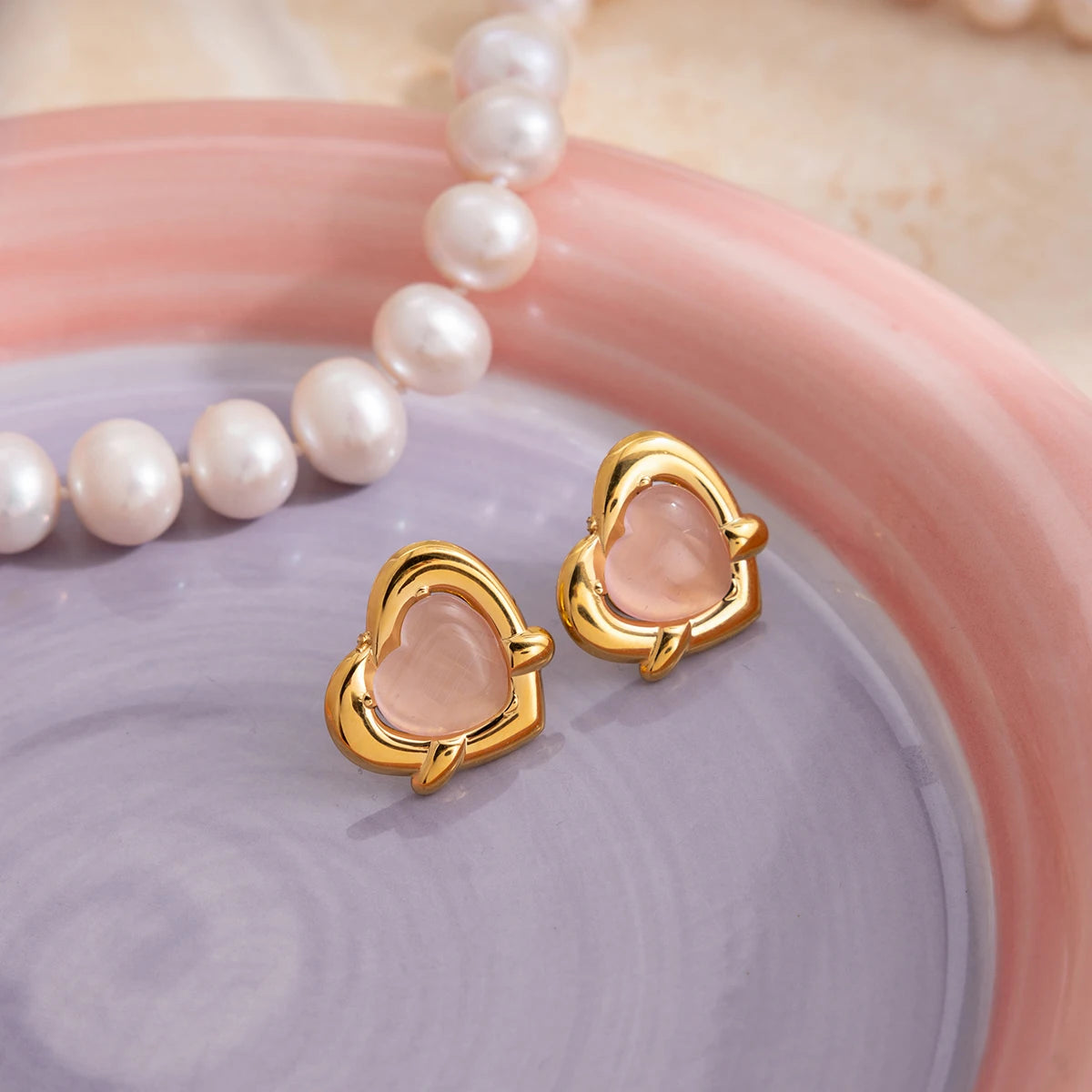 Stilvolle Produktfotografie der goldenen Herz-Ohrstecker mit rosa Cat-Eye-Stein auf einer bunten Schmuckschale neben einer Perlenkette.