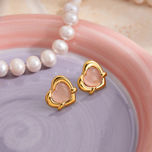 Stilvolle Produktfotografie der goldenen Herz-Ohrstecker mit rosa Cat-Eye-Stein auf einer bunten Schmuckschale neben einer Perlenkette.