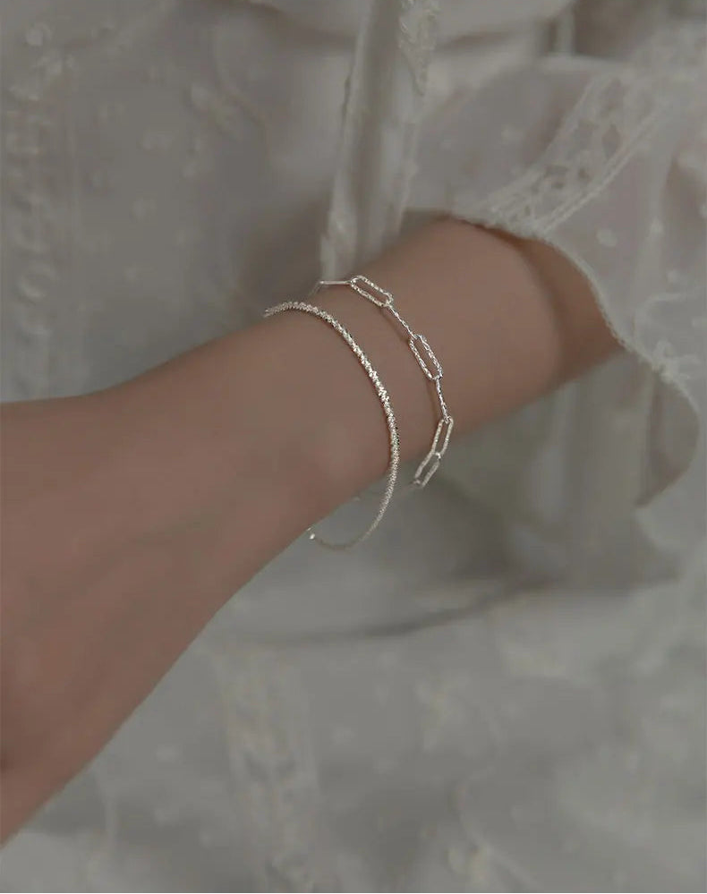  Handgelenk, das zwei zarte, silberfarbene Armbänder trägt (ein Gliederketten-Armband und ein dünnes Kettchen), vor dem Hintergrund eines weißen, rüschenbesetzten Oberteils.

